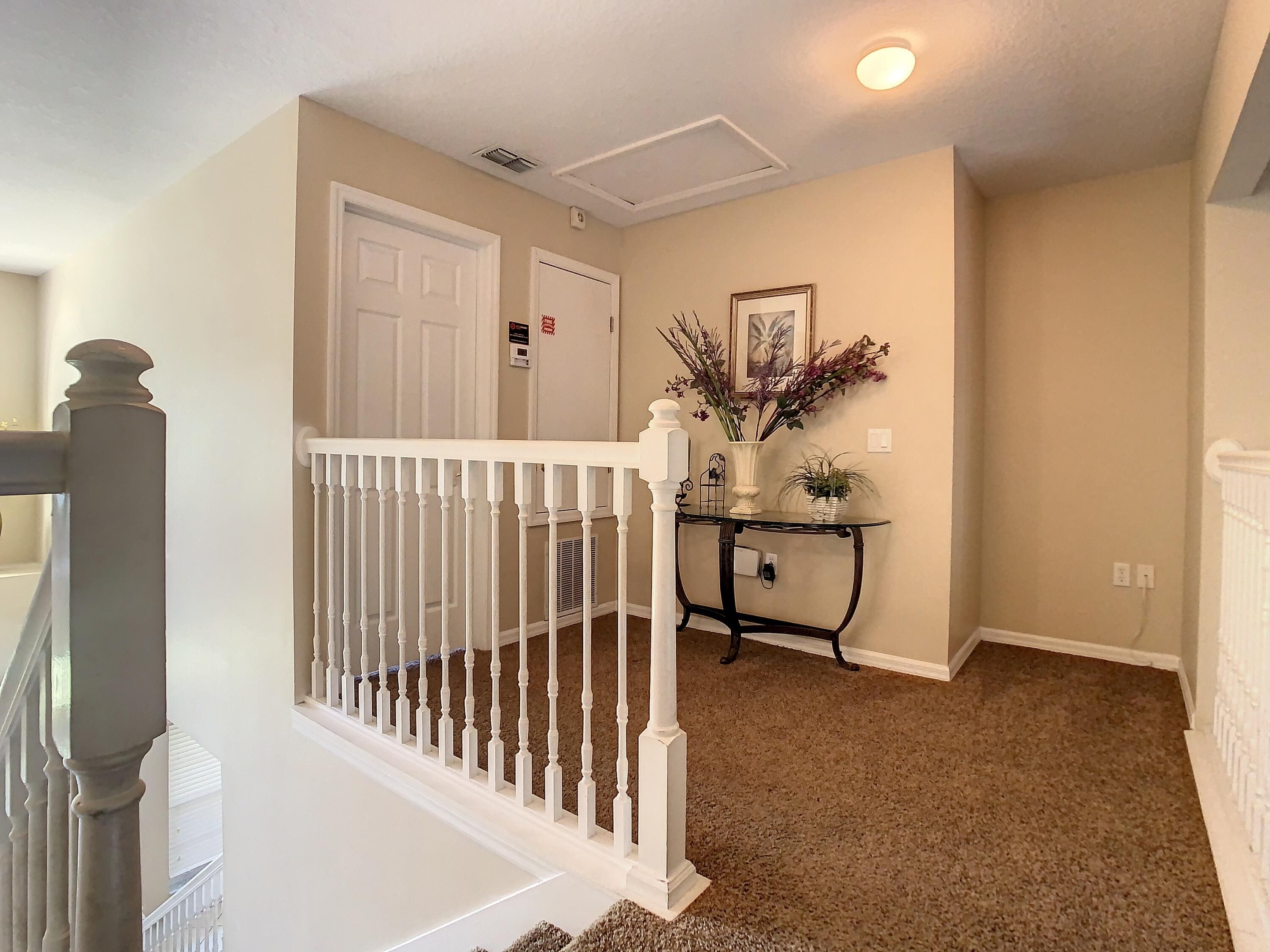 A hallway with a white door, a small table with a vase and plants, and a carpeted floor.