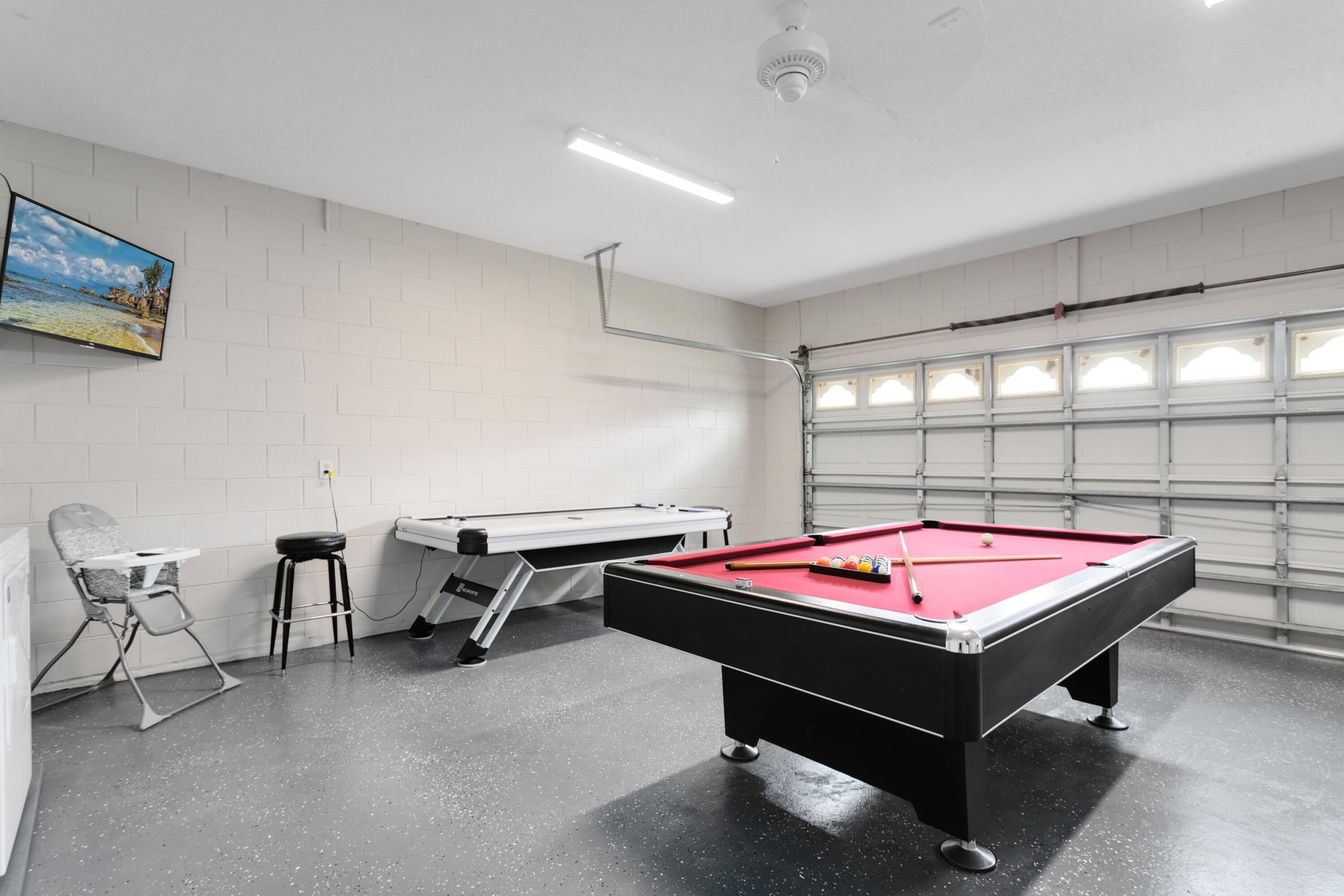 A pool table with a red felt surface, a white pool table, and a pool cue in a room with white brick walls and a large window.