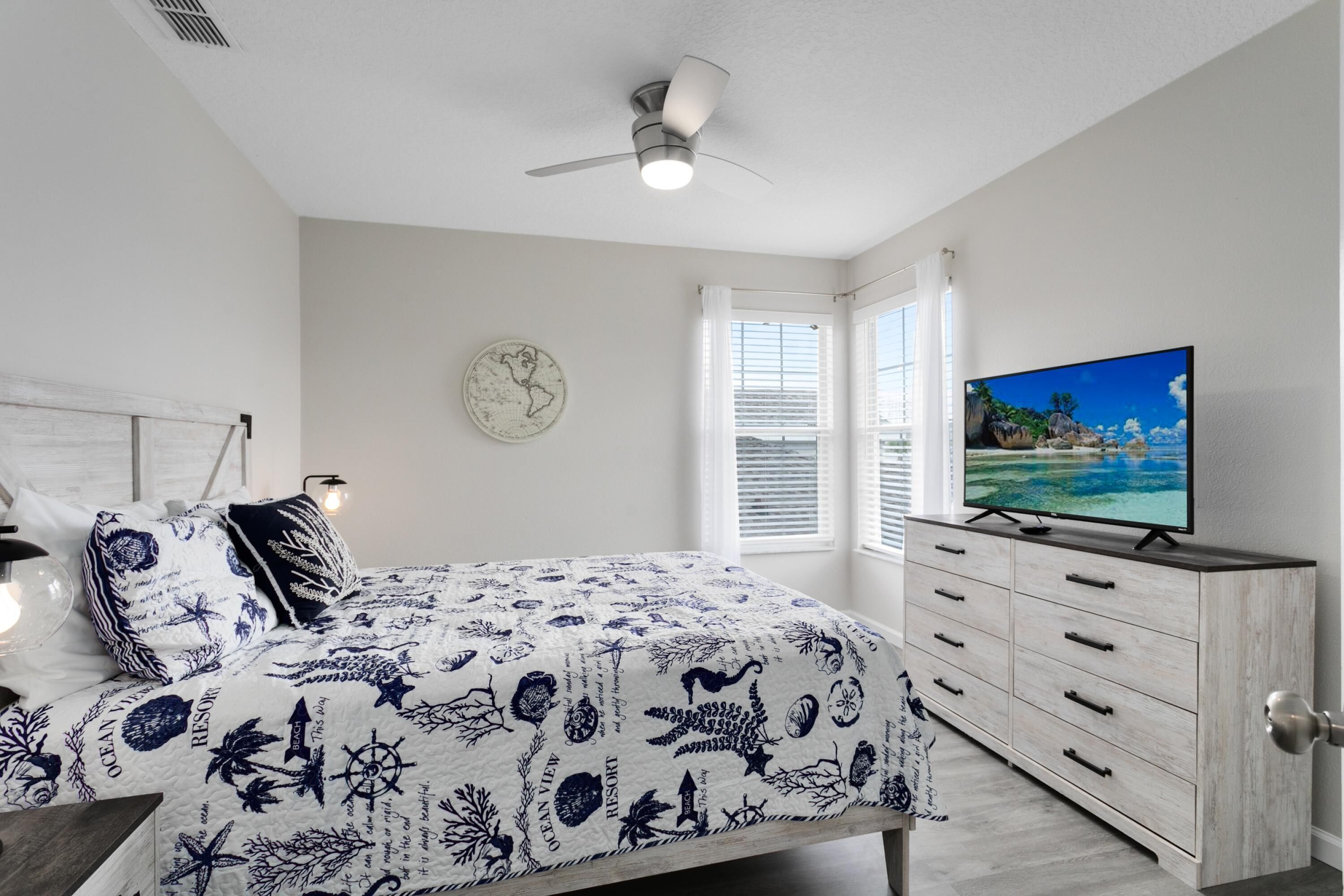 A bedroom with a bed, a TV, a dresser, a ceiling fan, a window, and a wall clock.