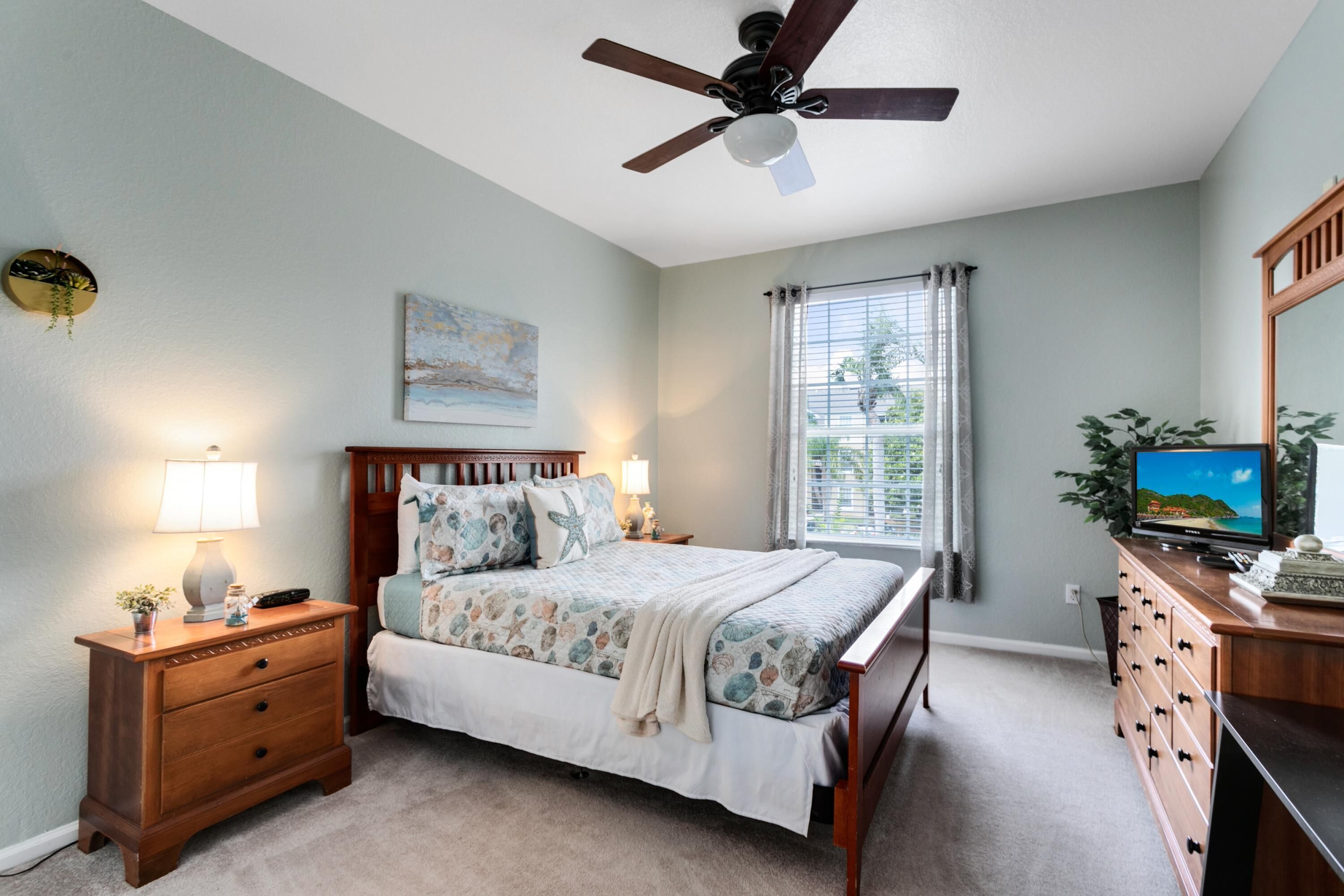 Une chambre à coucher comprenant un lit, une table de chevet, une commode, un ventilateur de plafond, une fenêtre avec des rideaux et un téléviseur.