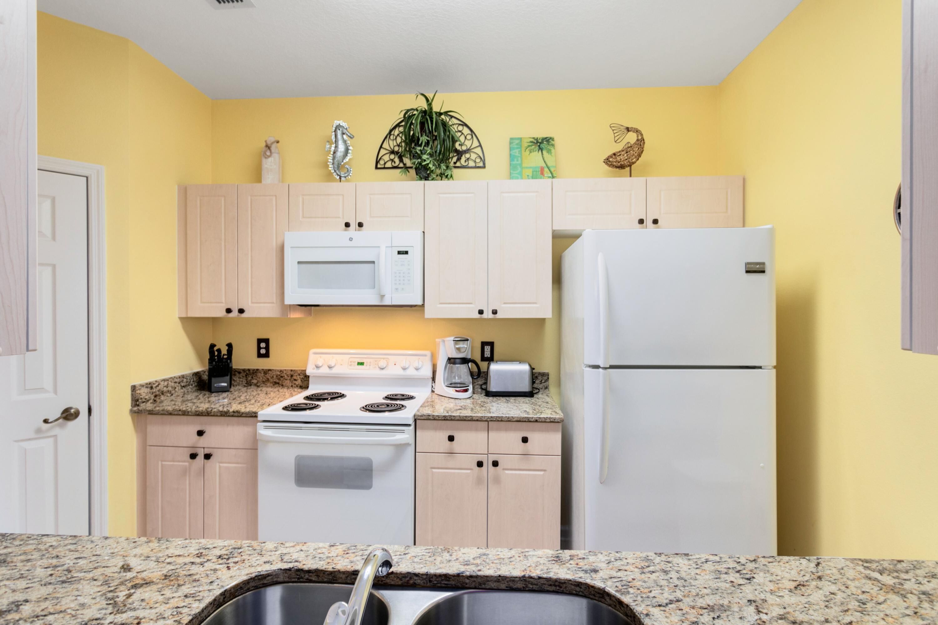 Une cuisine compacte dotée d’une cuisinière blanche, d’un four à micro-ondes et d’un réfrigérateur, de meubles de rangement en bois clair, de plans de travail en granit et d’un évier.
