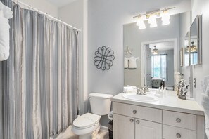 A bathroom with a striped shower curtain, a white toilet, a sink with a mirror, and decorative wall art. - Close to Disney Condo 3 Bed 2 Bath (Kissimmee)