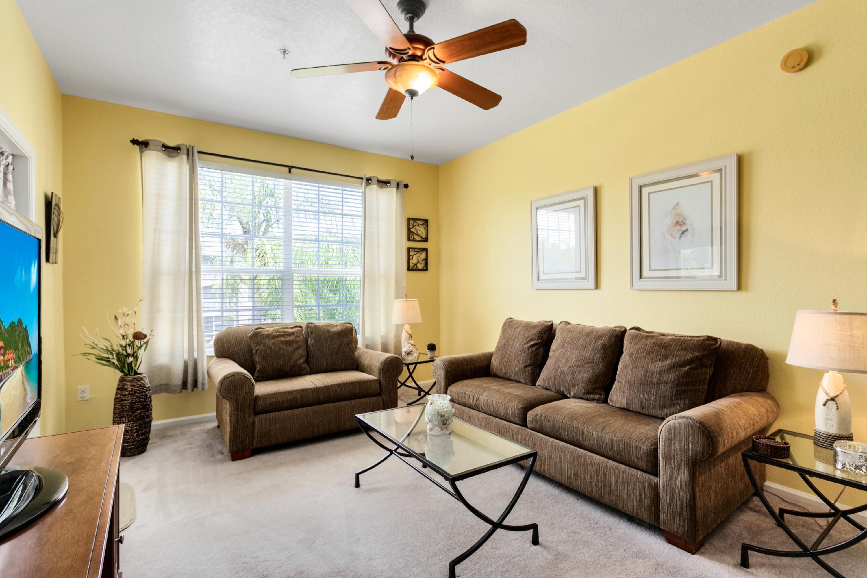 A living room with two brown sofas, a glass coffee table, a ceiling fan, and a TV on a wooden stand.