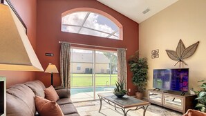 A living room with a leather sofa, a coffee table, a TV, and a large window overlooking a backyard. - Emerald Island Resort 4 bed 3 Bath With Pool spa (Kissimmee)