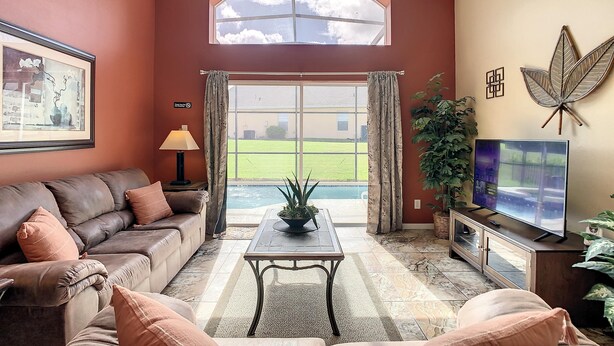 A living room with a brown leather couch, a coffee table, a TV, and a large window with curtains. - Emerald Island Resort 4 bed 3 Bath With Pool spa (Kissimmee)