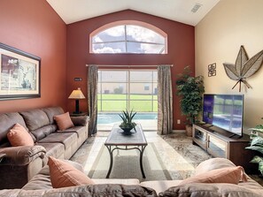 A living room with a brown leather couch, a coffee table, a TV, and a large window with curtains.