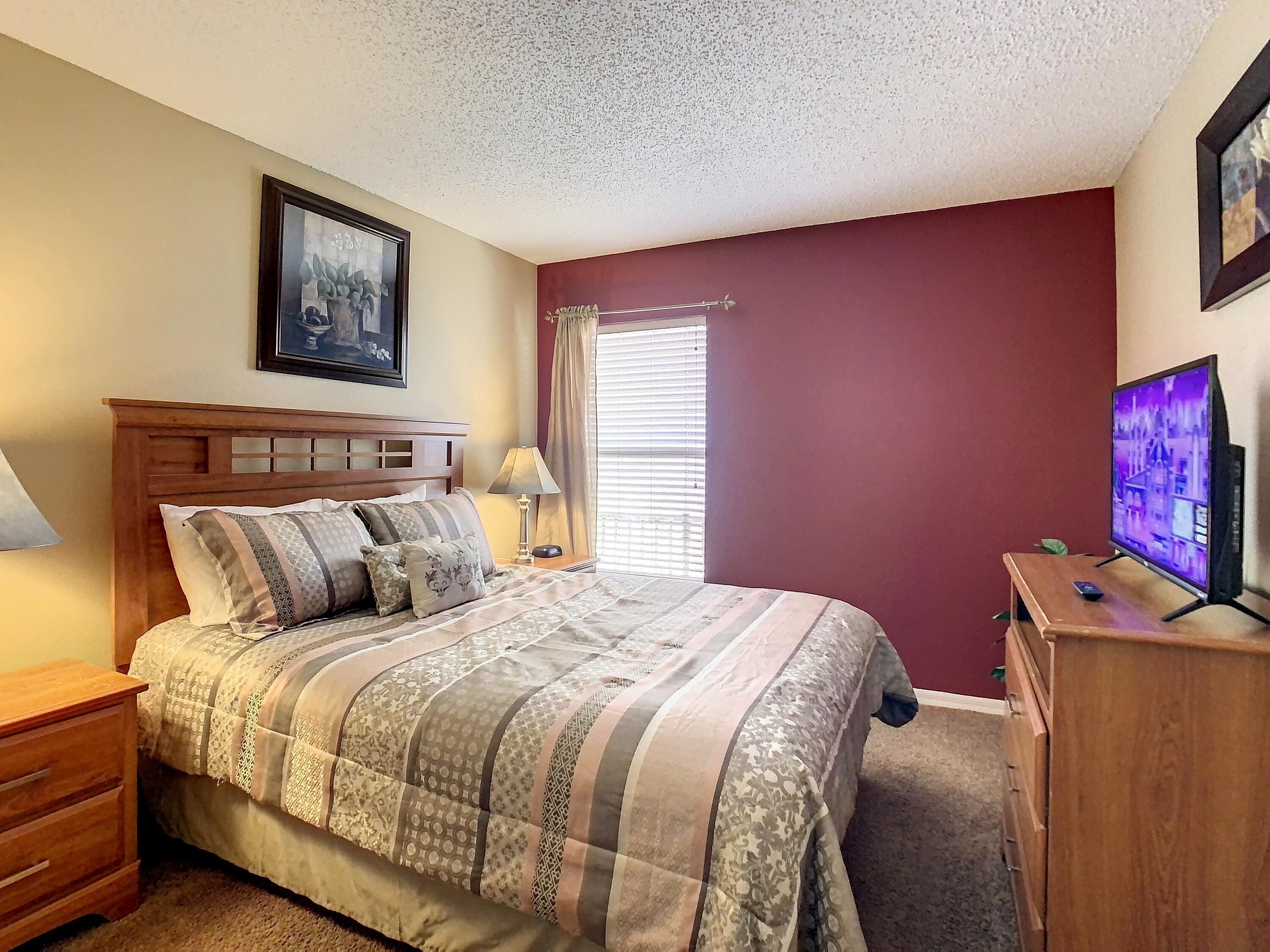 A bedroom with a wooden bed, a nightstand, a dresser, a TV, and a window with blinds.