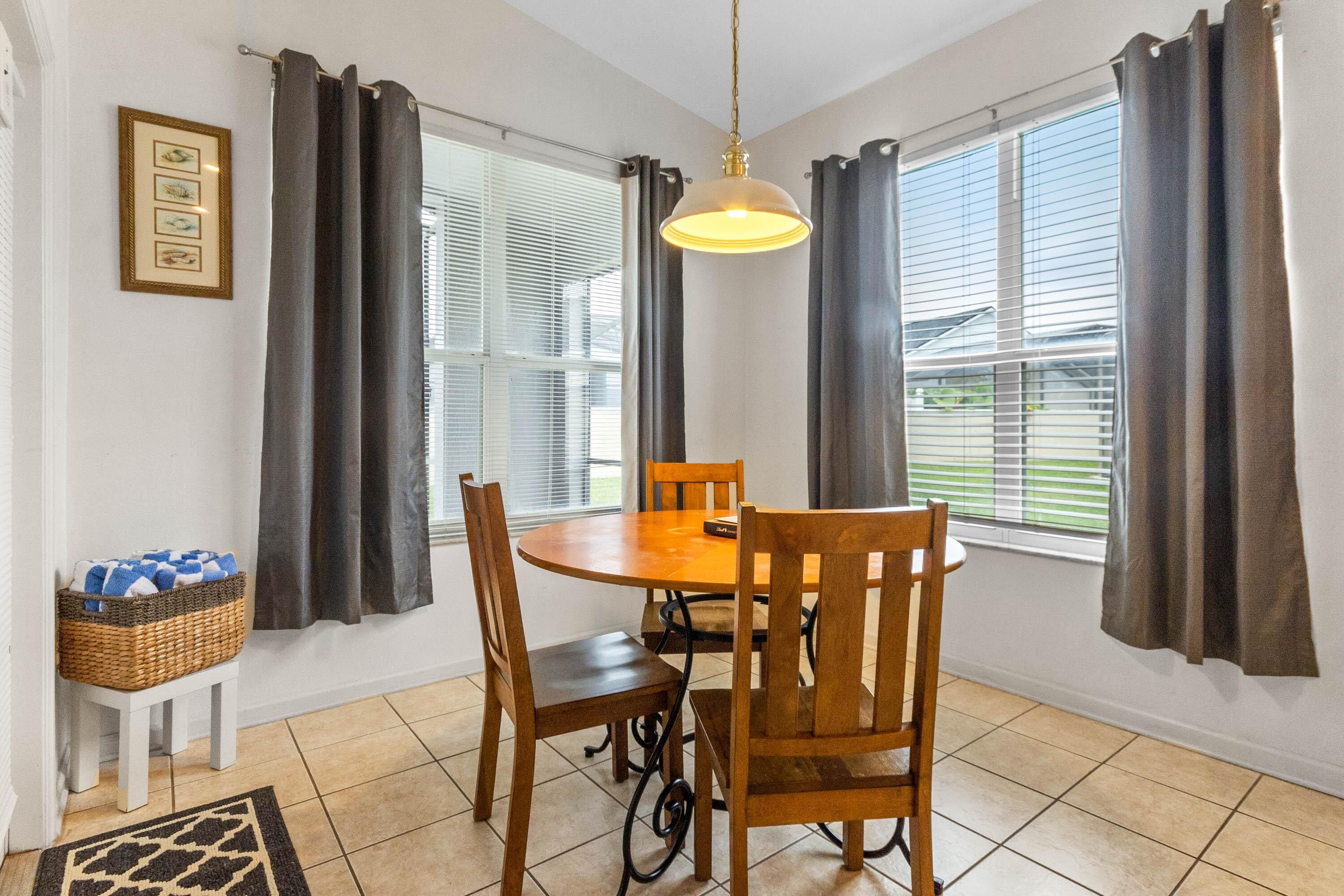A dining area with a round wooden table and four chairs, a hanging pendant light, and a window with blinds.