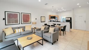 A modern living room with a sofa, armchairs, and a coffee table. There is a dining area with a table and chairs. The kitchen is visible in the background with cabinets and a refrigerator. - Amazing Townhouse Close to Disney 2 Bed 2 5 Bath (Kissimmee)