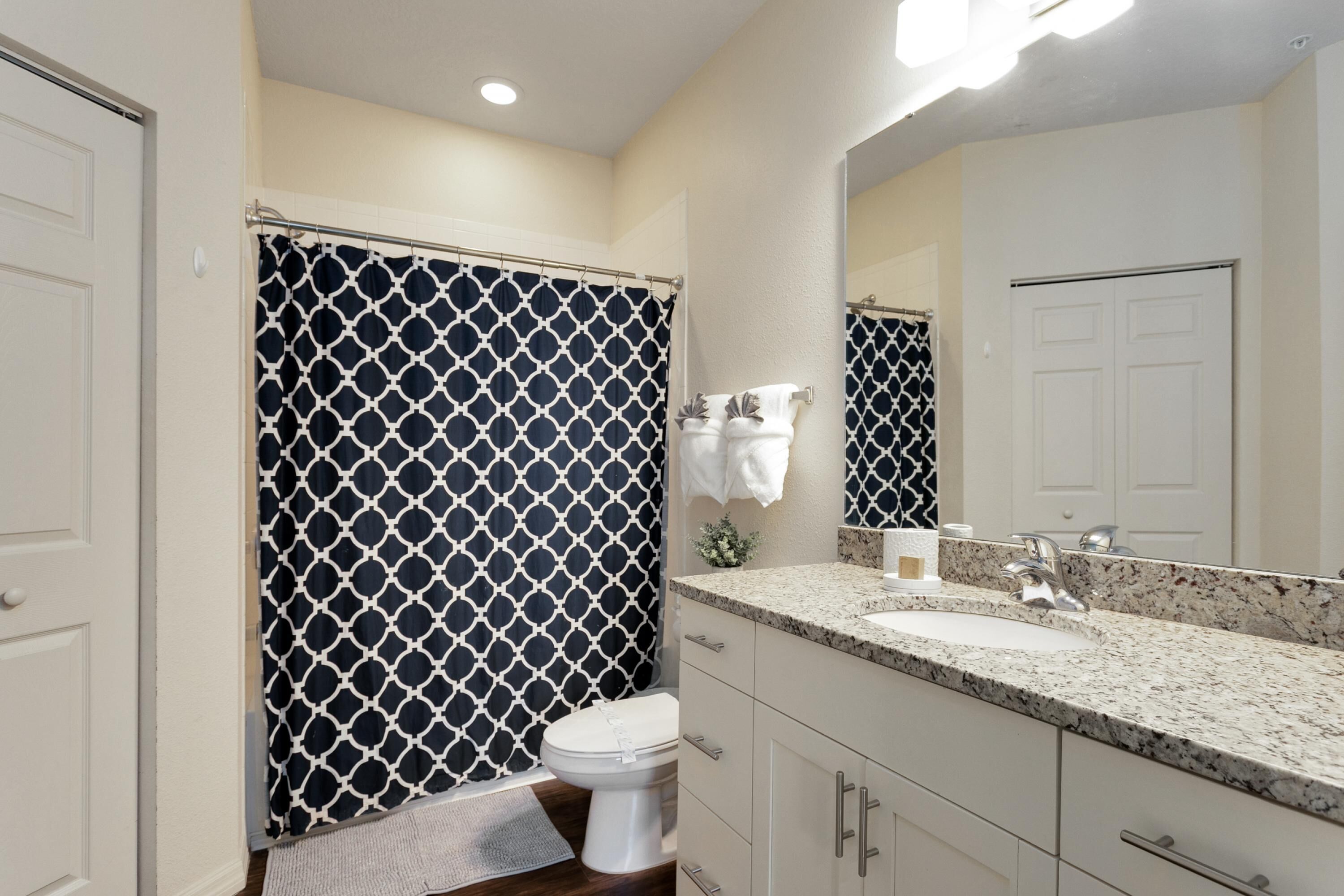 Un baño con cortina de ducha estampada, un inodoro blanco, un lavabo con encimera de granito y un espejo sobre el lavabo.
