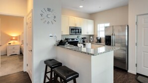 A modern kitchen with white cabinets, a stainless steel refrigerator, a microwave, and a granite countertop. There are two bar stools and a clock on the wall. - Lucaya Village Resort - 4 Bed 3 Bath Townhouse (Kissimmee)