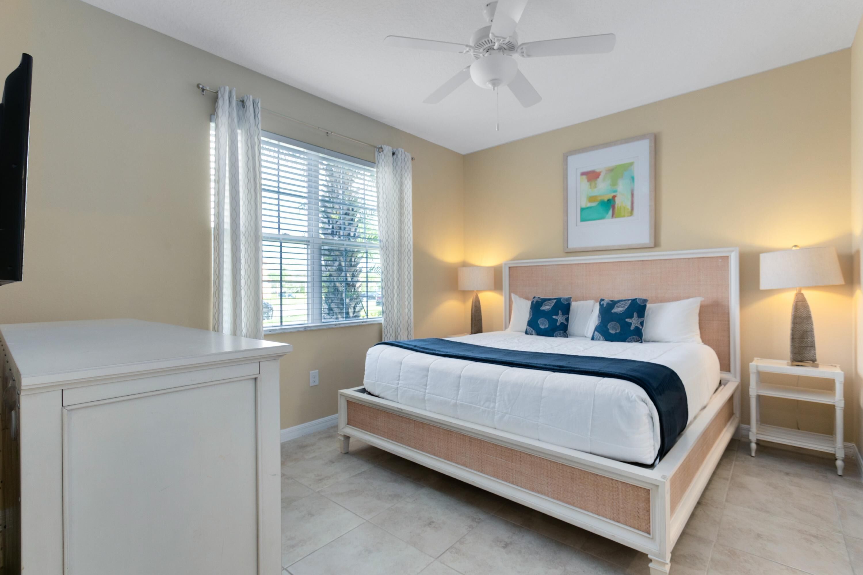 Une chambre à coucher comprenant un lit, une table de chevet, un ventilateur de plafond, une fenêtre avec des rideaux et une œuvre d’art accrochée au mur.