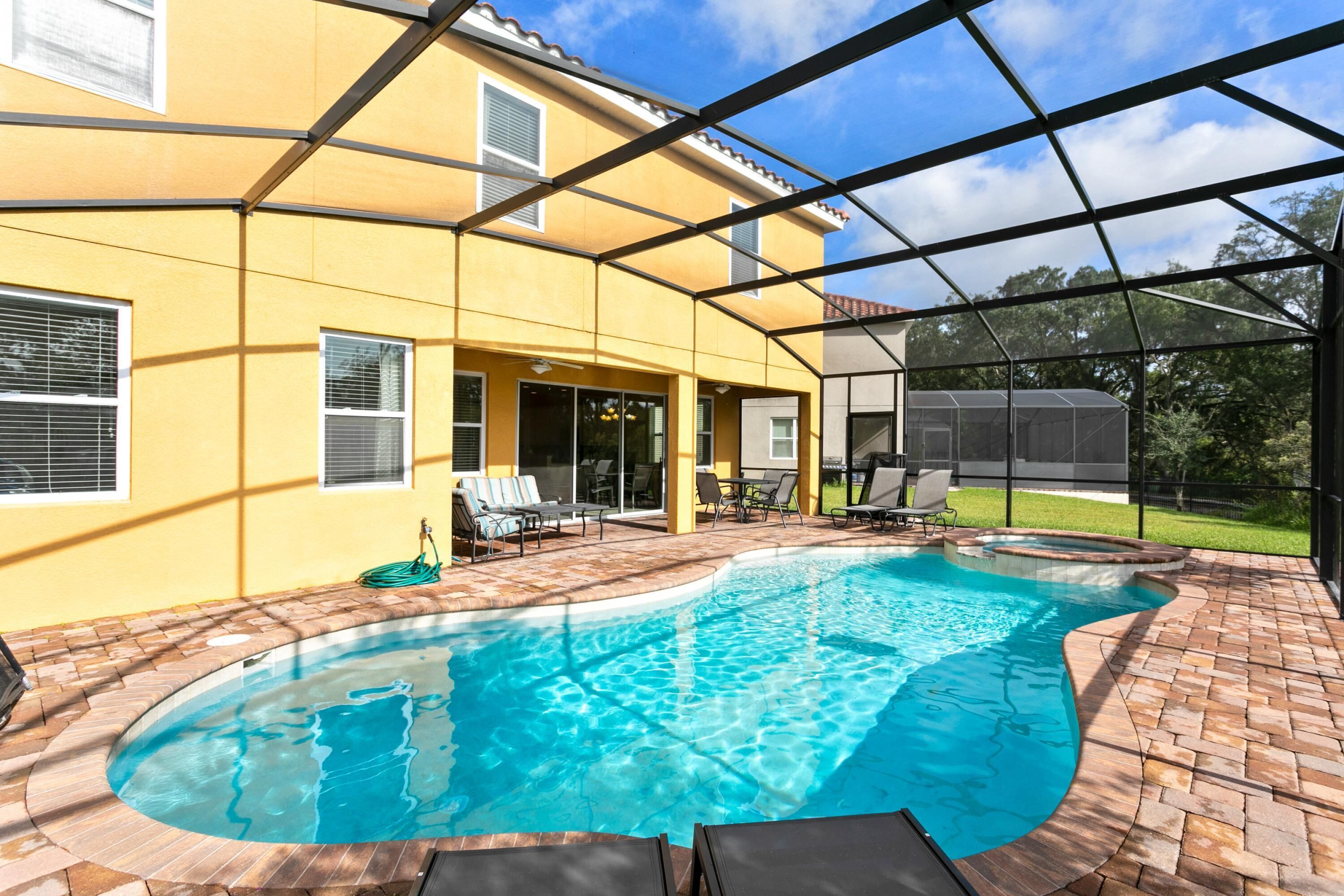 A swimming pool with a brick border, a covered patio area, and a yellow building with windows and a door.