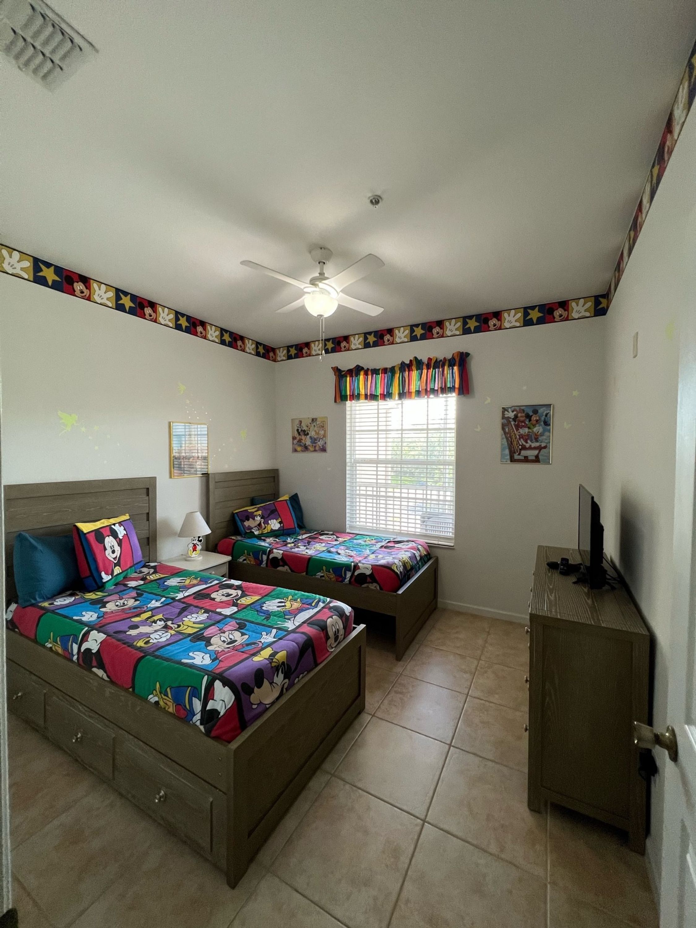 A bedroom with two beds, a ceiling fan, a window with blinds, a TV on a wooden cabinet, and a Mickey Mouse themed border on the walls.