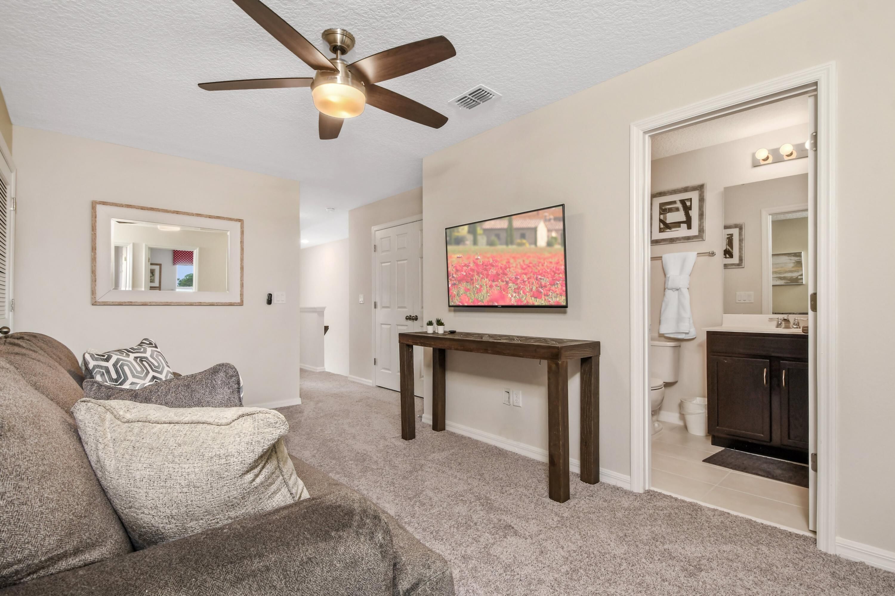 A living room with a ceiling fan, a large flat-screen TV, a dark wooden coffee table, and a beige carpet.