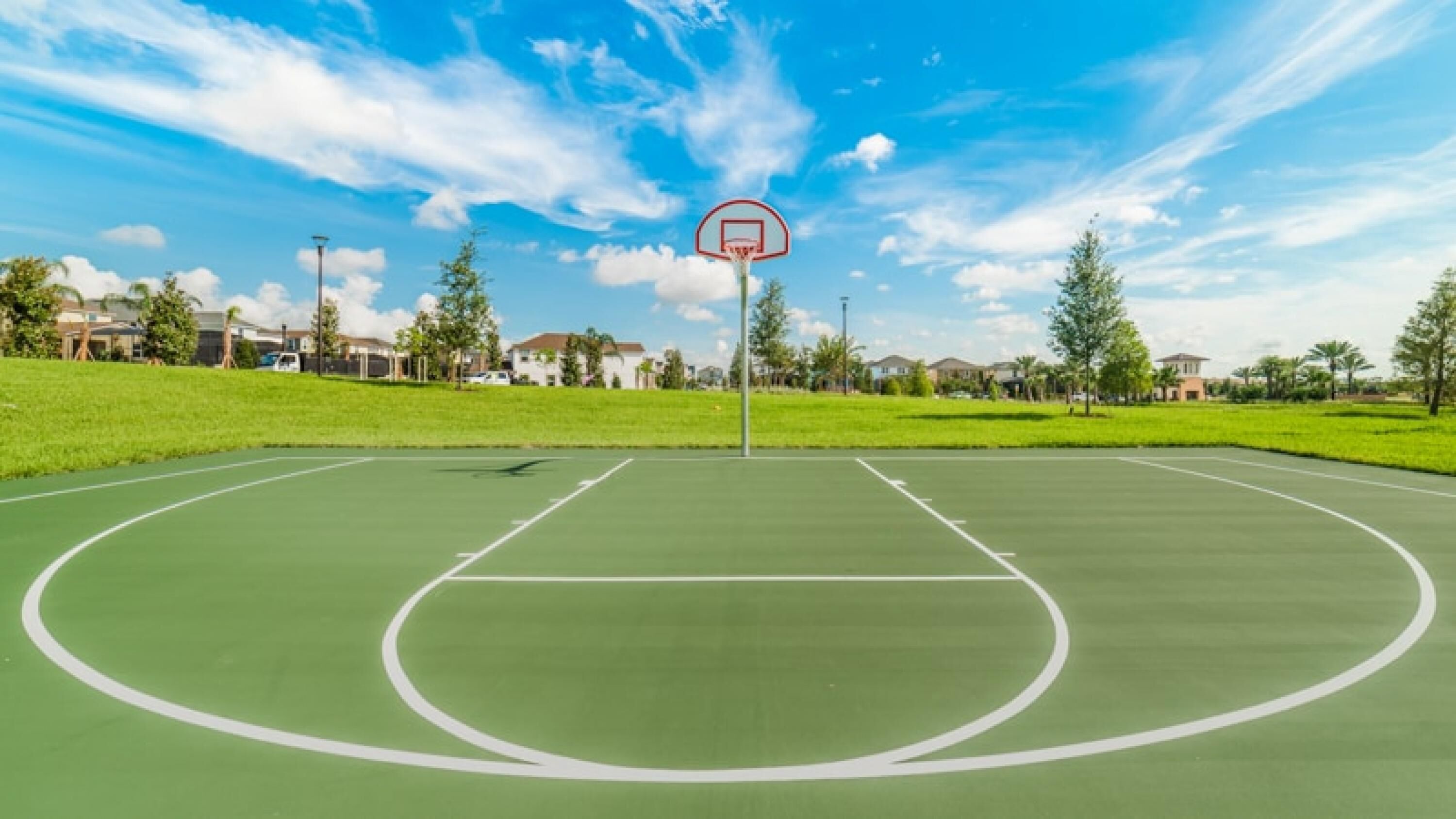 An outdoor basketball court with a clear sky, surrounded by green grass and trees.