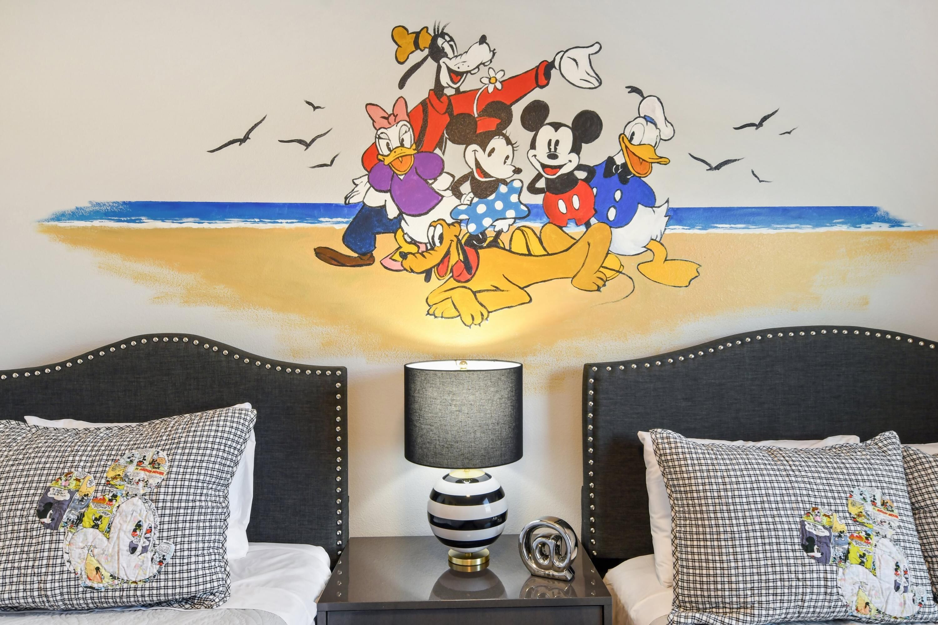 Habitación de hotel con una cama, dos mesitas de noche, una lámpara y un mural en la pared con personajes animados.