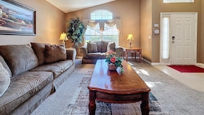 A spacious living room with a large sofa, a coffee table, and two armchairs. There is a large window with curtains, a door, and a framed picture on the wall.