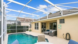 Un espace extérieur abrité avec une piscine, comprenant une table et des chaises blanches, un canapé et un tuyau d’arrosage fixé au mur.