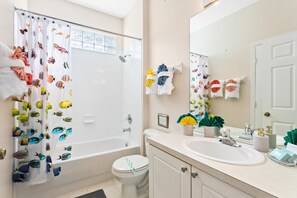 Een badkamer met een bad, toilet en wastafel. Handdoeken hangen aan de muur en een douchegordijn met vismotieven.