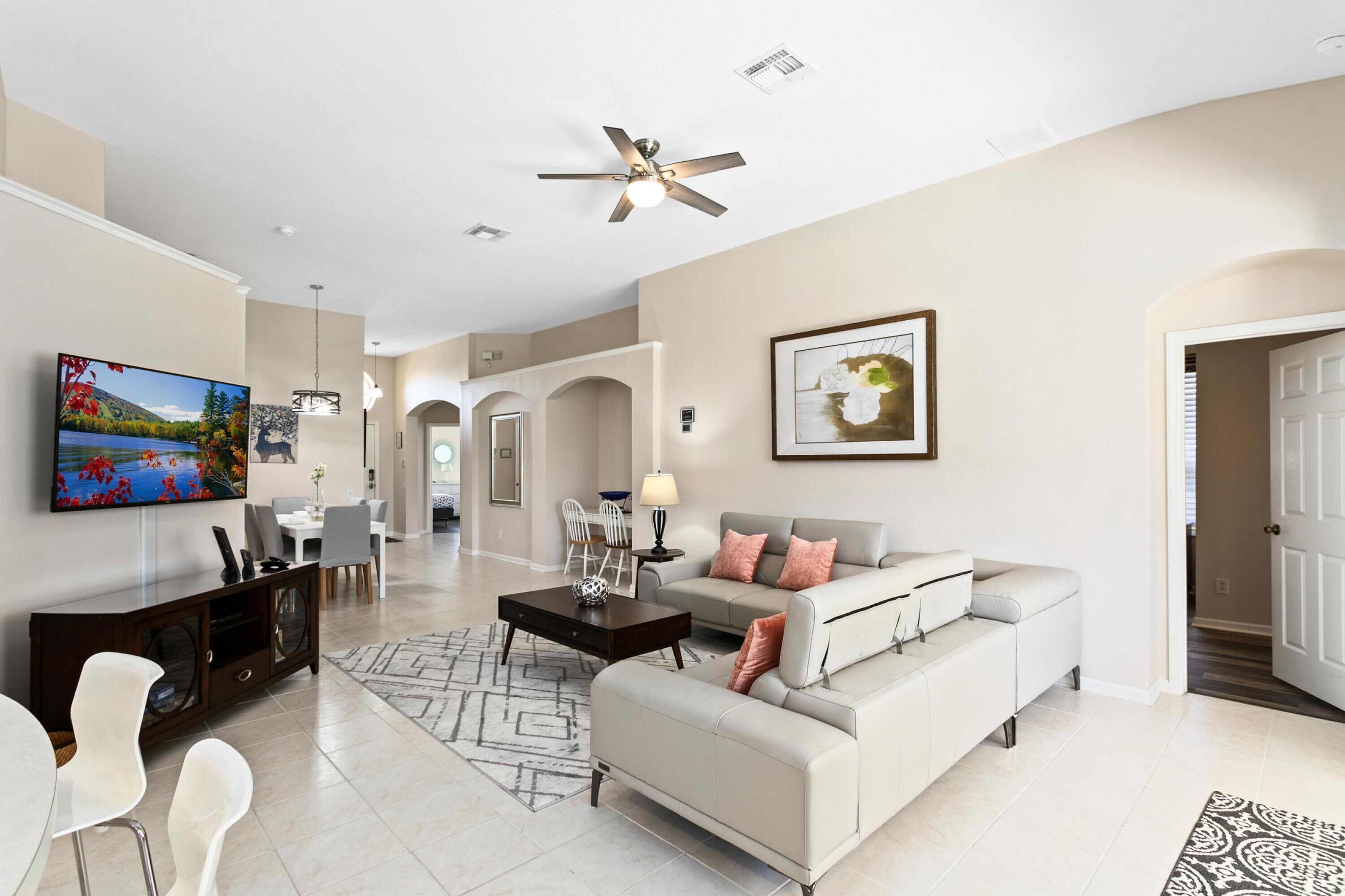 A spacious living room with a large flat-screen TV, a ceiling fan, and a sofa with cushions.