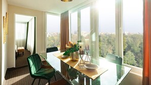 A modern hotel room with a dining area featuring a glass table, a green upholstered chair, and a view of a forested landscape through large windows. - Qult·co - The Renaissance Affair (București)