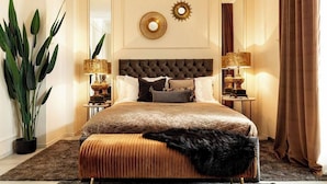 A bedroom with a tufted headboard, a large bed with a quilted cover, two bedside lamps, a large potted plant, and a mirror on the wall. - Qult·co - The Aristocrat (București)