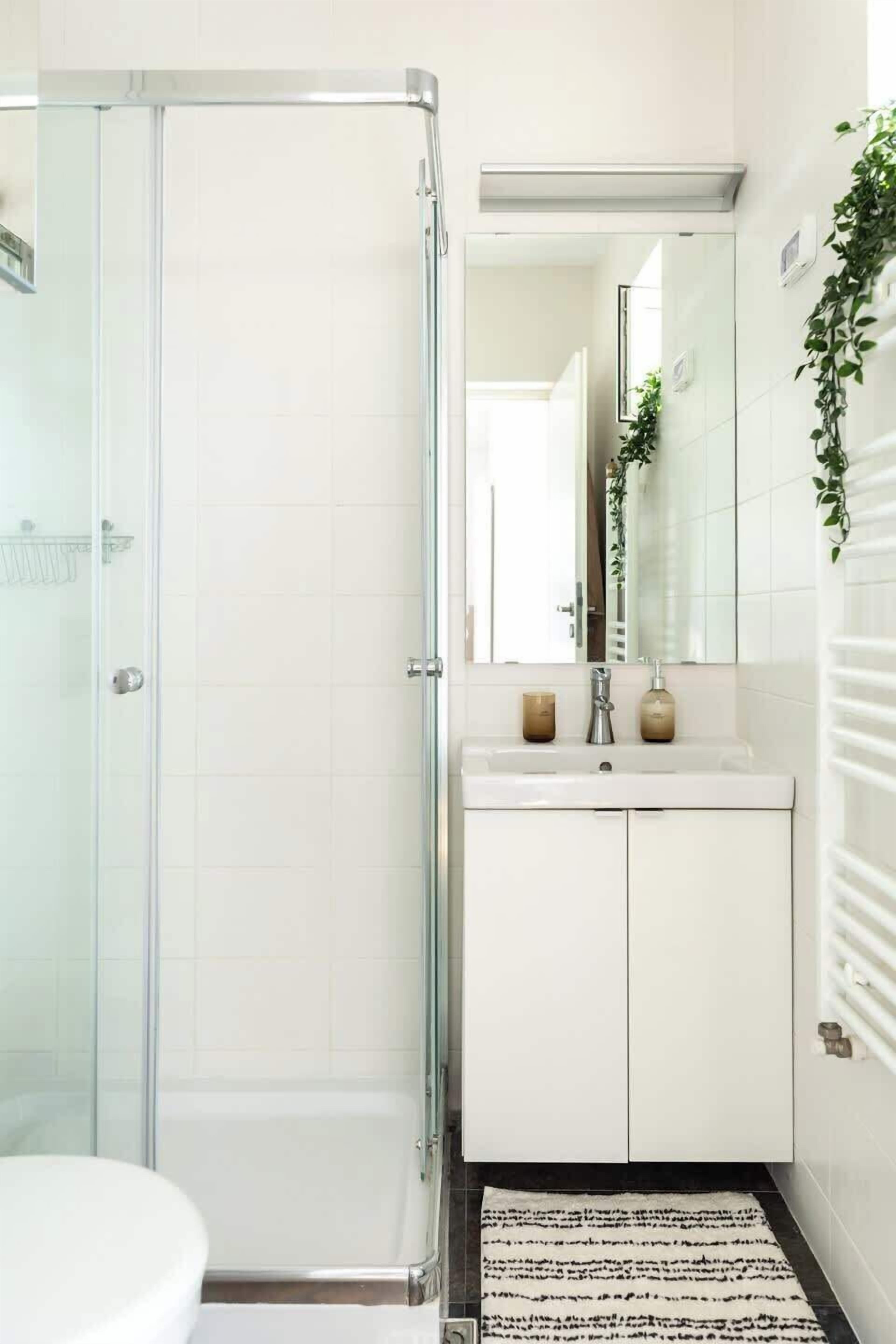 A modern bathroom with a glass shower enclosure, a white sink with a mirror above, and a patterned rug on the floor.
