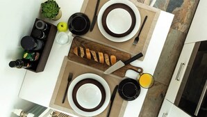 A top-down view of a kitchen with a dining table set for two, featuring plates, cutlery, a loaf of bread, a cutting board, a knife, a glass of orange juice, and a small potted plant.