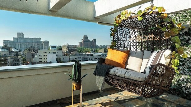 A balcony with a wicker sofa, a potted plant, and a view of city buildings.