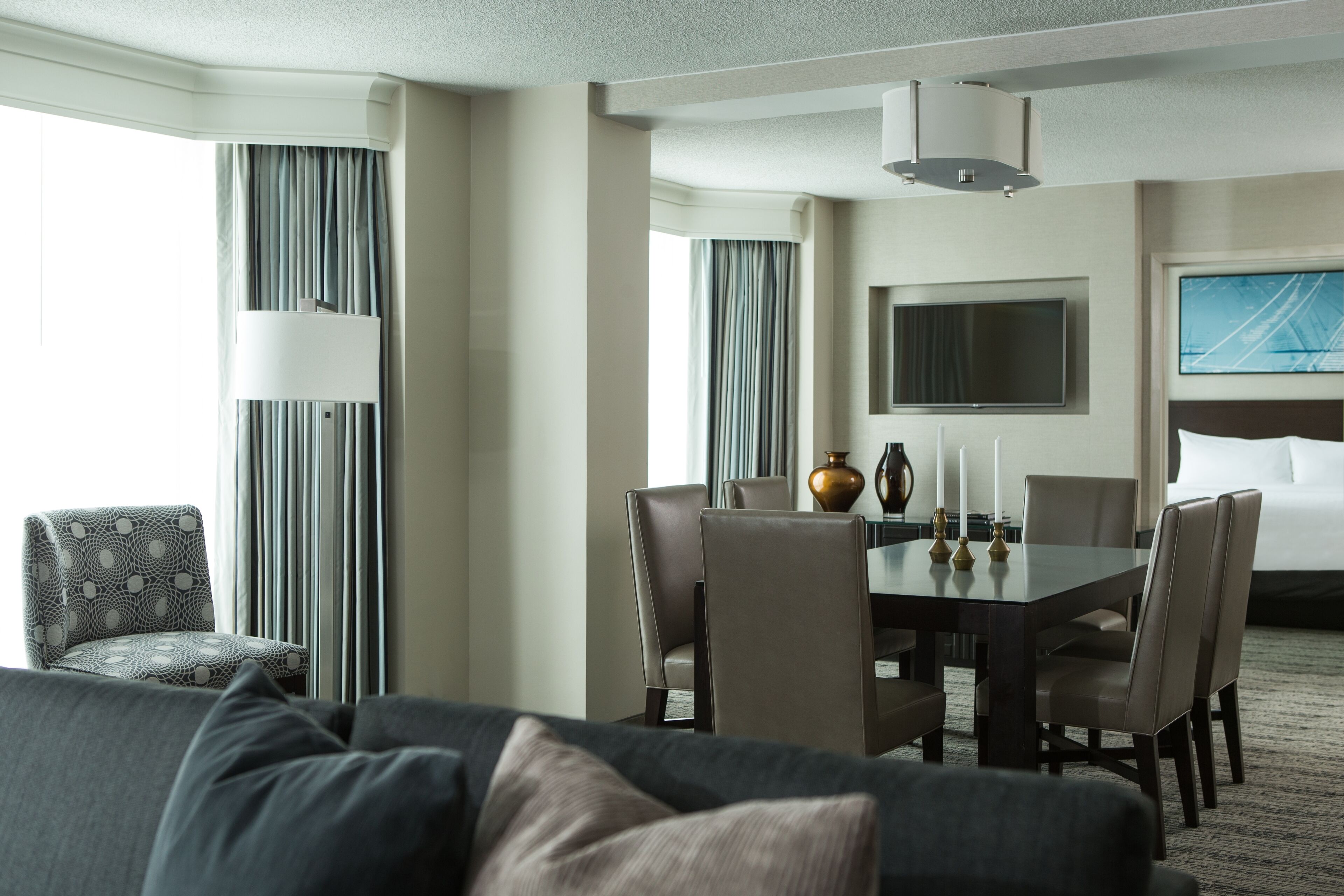 A hotel room with a dining area, a sofa, a TV, and a large window with curtains.