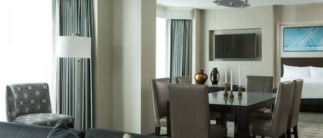 A hotel room with a dining area, a sofa, a TV, and a large window with curtains.