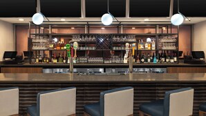 A bar area with a counter, shelves of bottles, glasses, and a cash register. There are three chairs in front of the bar.