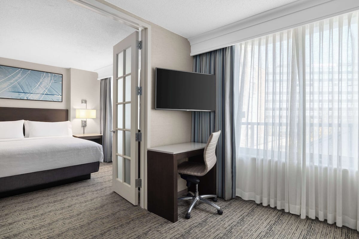 Ein Hotelzimmer mit Bett, Schreibtisch, Stuhl, Fernseher und großem Fenster mit Vorhängen.
