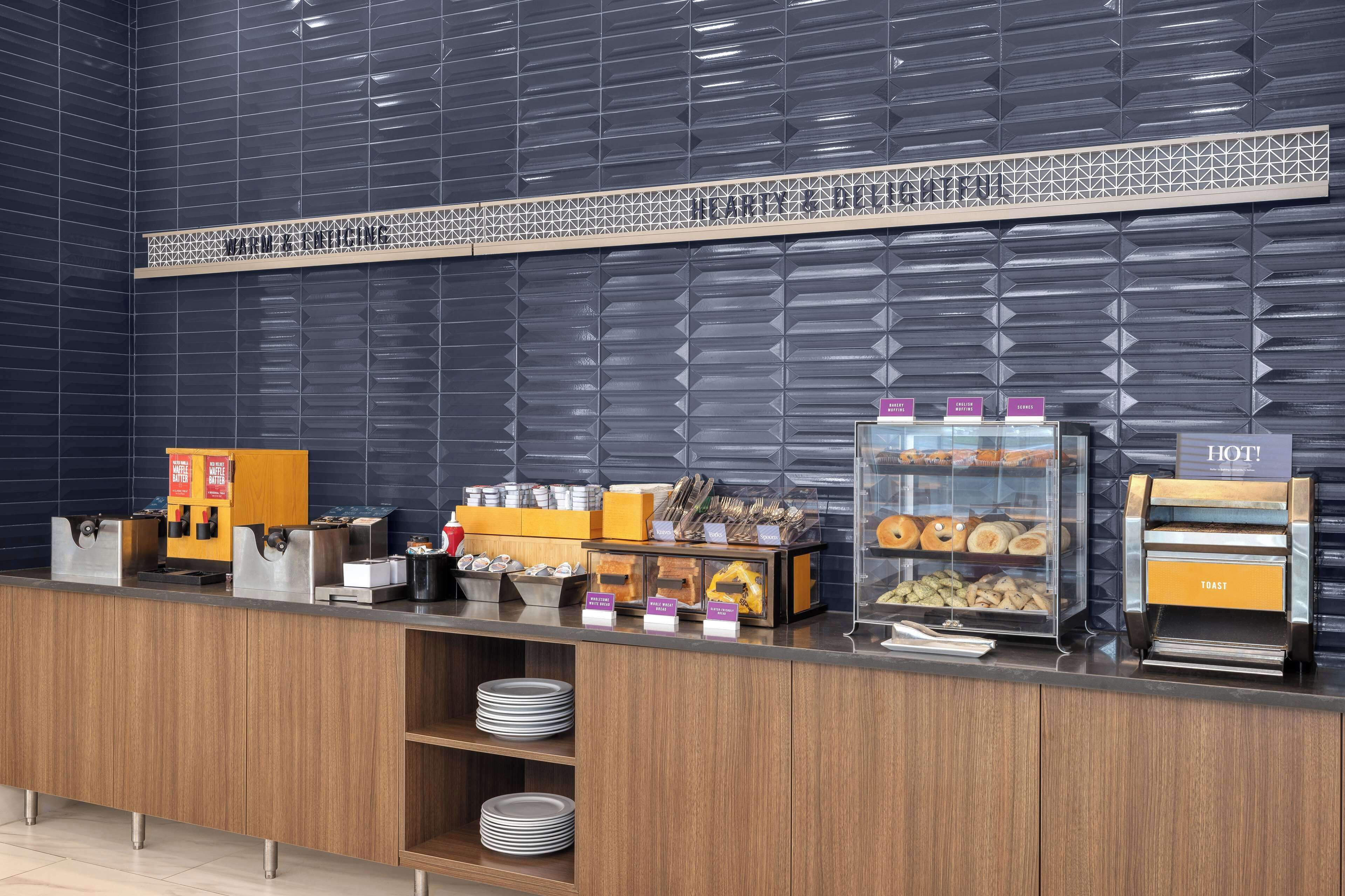 Zona de desayuno de hotel con un mostrador que exhibe diferentes alimentos y una máquina de café.