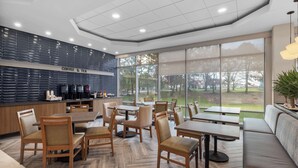 A modern cafe with wooden furniture, a coffee bar, and large windows offering a view of the outdoors.