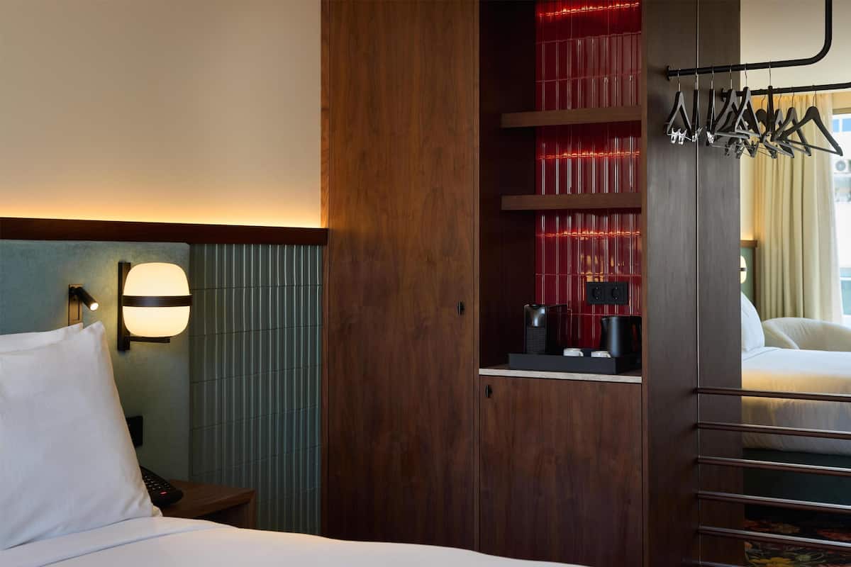 Une chambre d’hôtel avec un lit, une table de chevet, une lampe, une étagère éclairée en rouge et un porte-manteau.