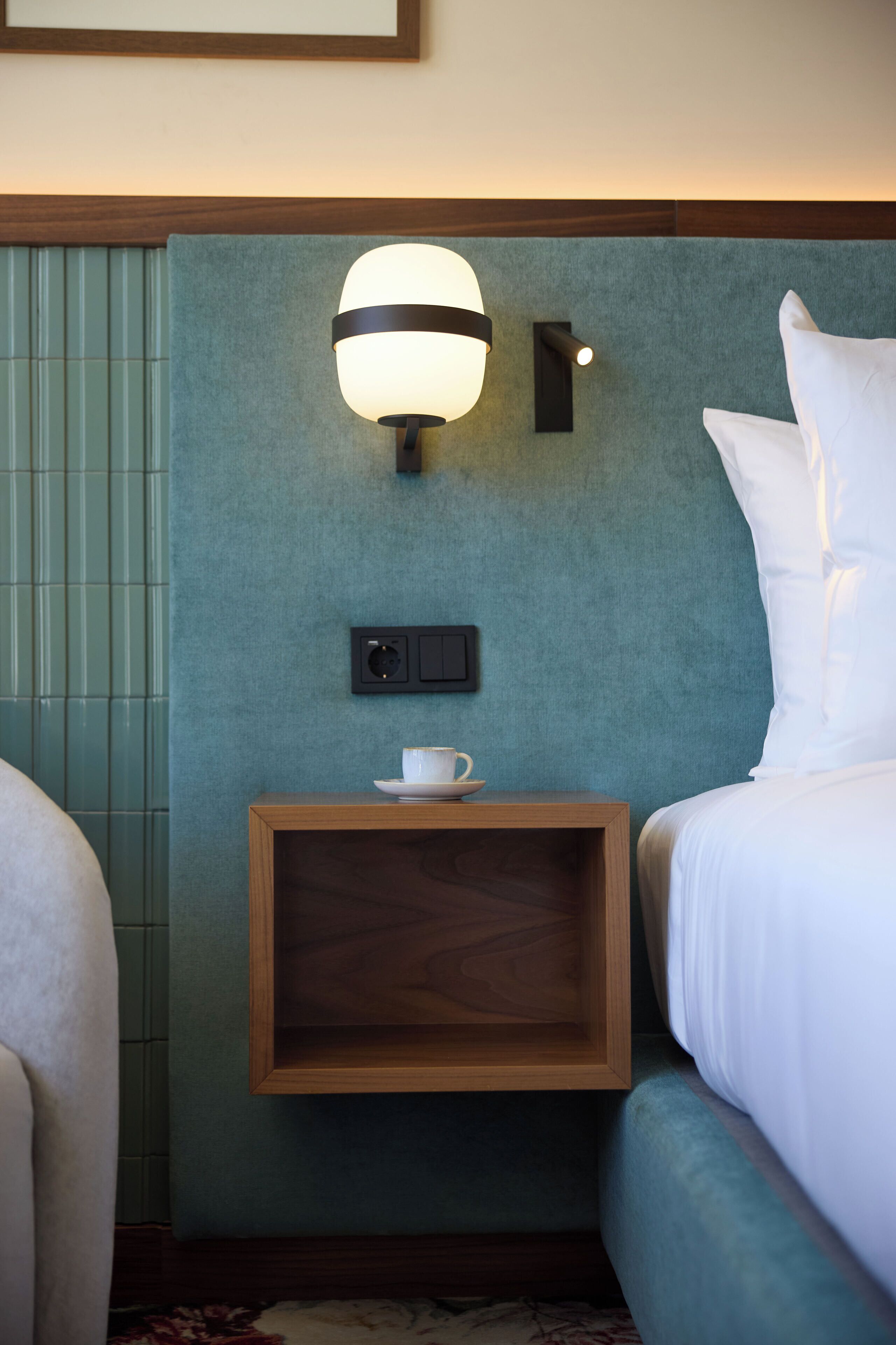 A hotel room with a bed, a nightstand with a lamp, and a small table with a cup.