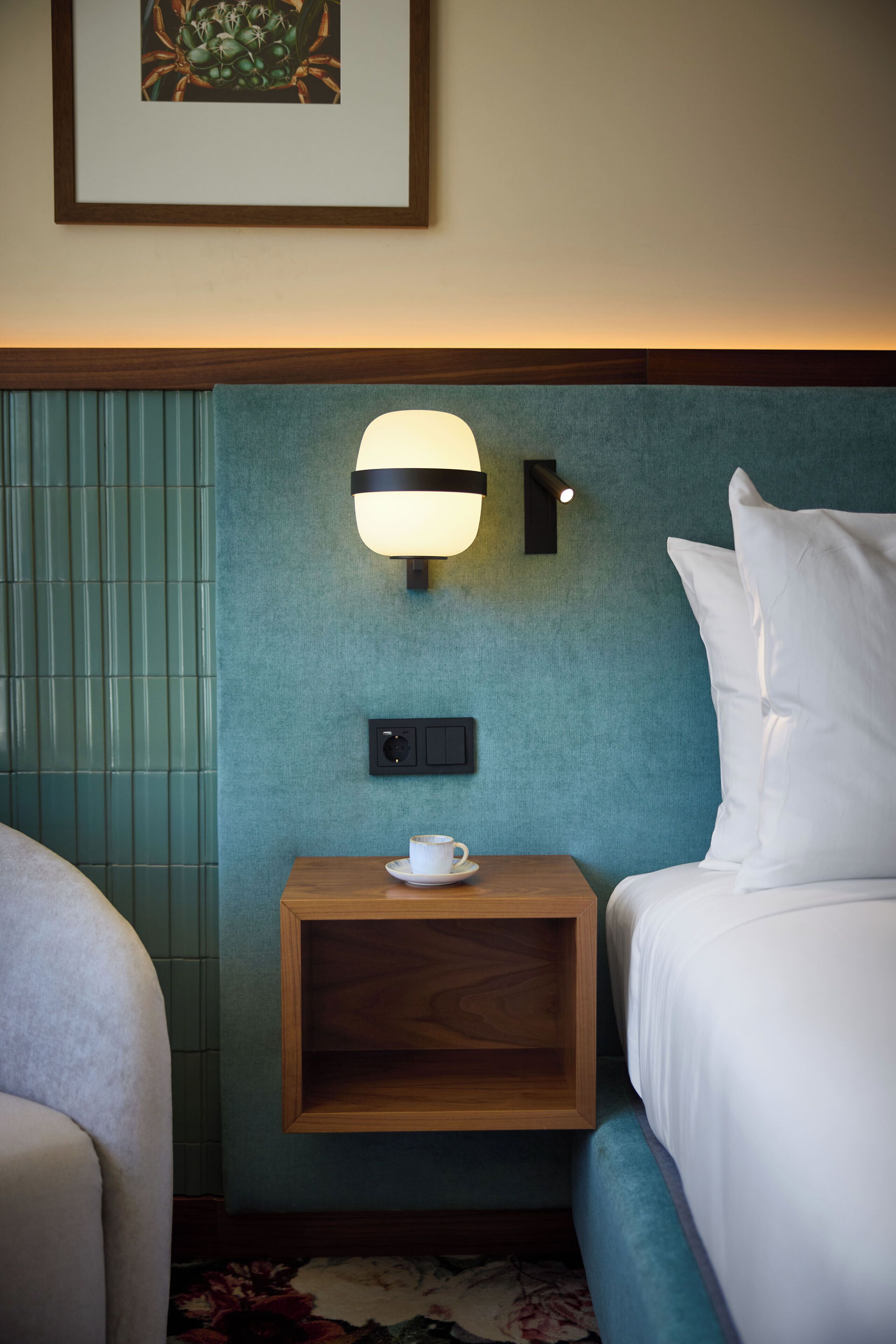 Una habitación de hotel con una cama, una mesita de noche con una taza y un plato, una lámpara de pared y un cuadro en la pared.