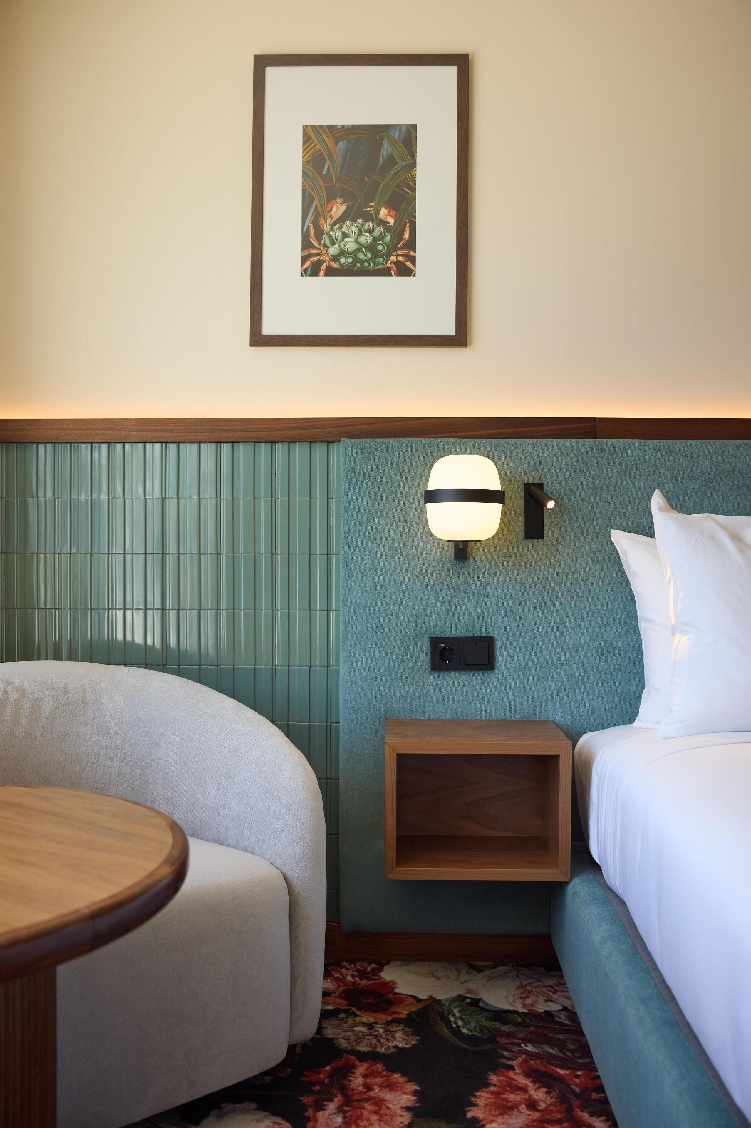 A hotel room with a bed, a chair, a small table, a wall-mounted light, and a framed picture.