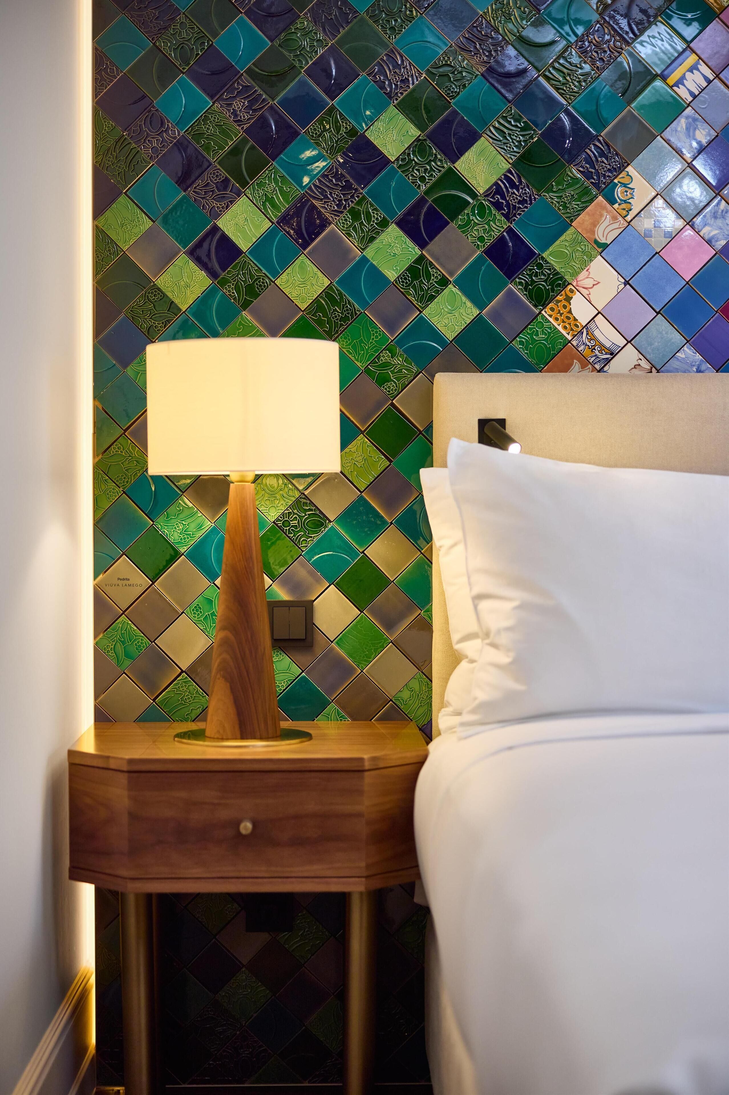 Una cama bien tendida con ropa blanca, una mesita de noche de madera con una lámpara, y una pared con un diseño de baldosas geométricas.
