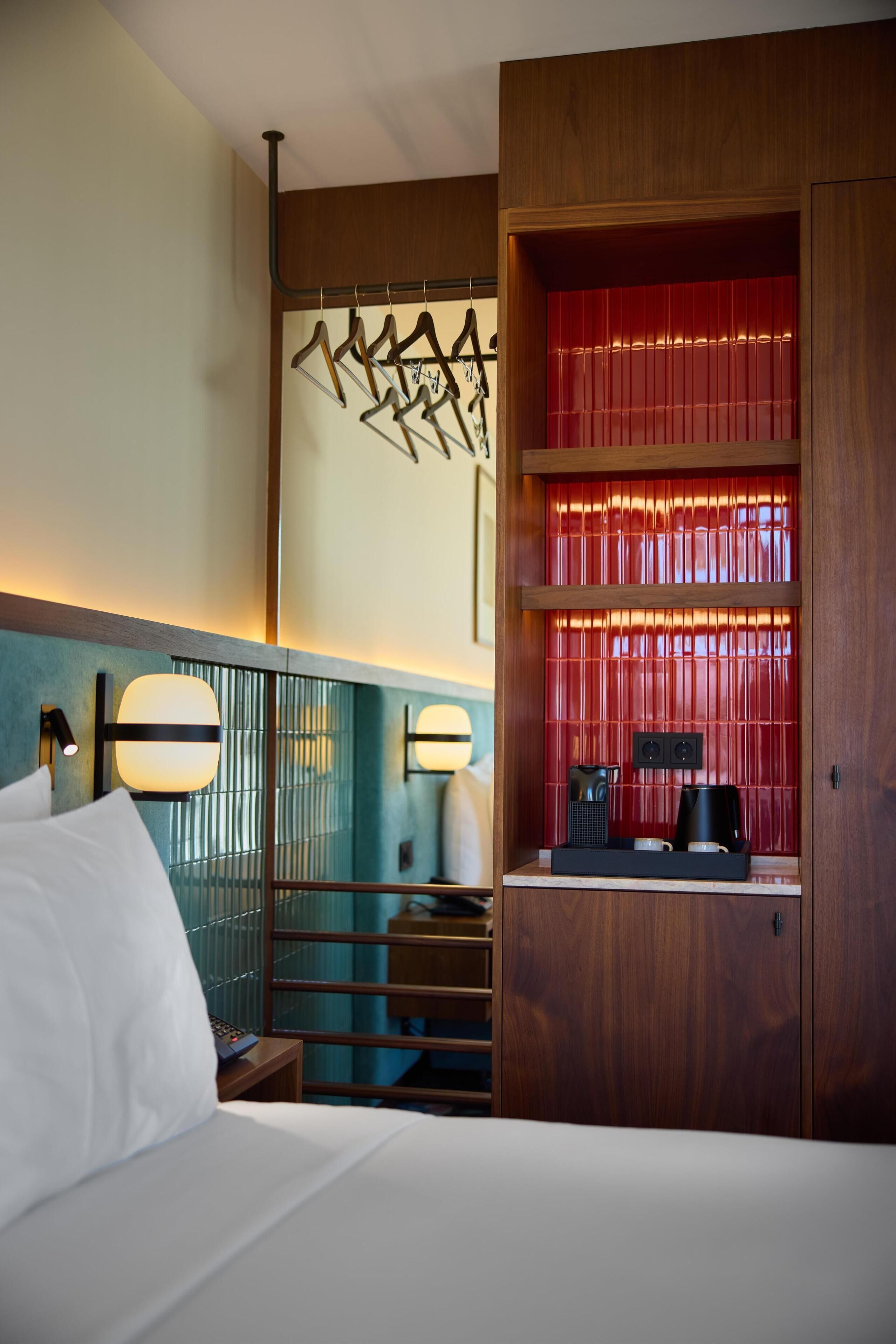 A hotel room with a bed, a nightstand, a wall-mounted lamp, a shelf with hanging items, and a red partition.