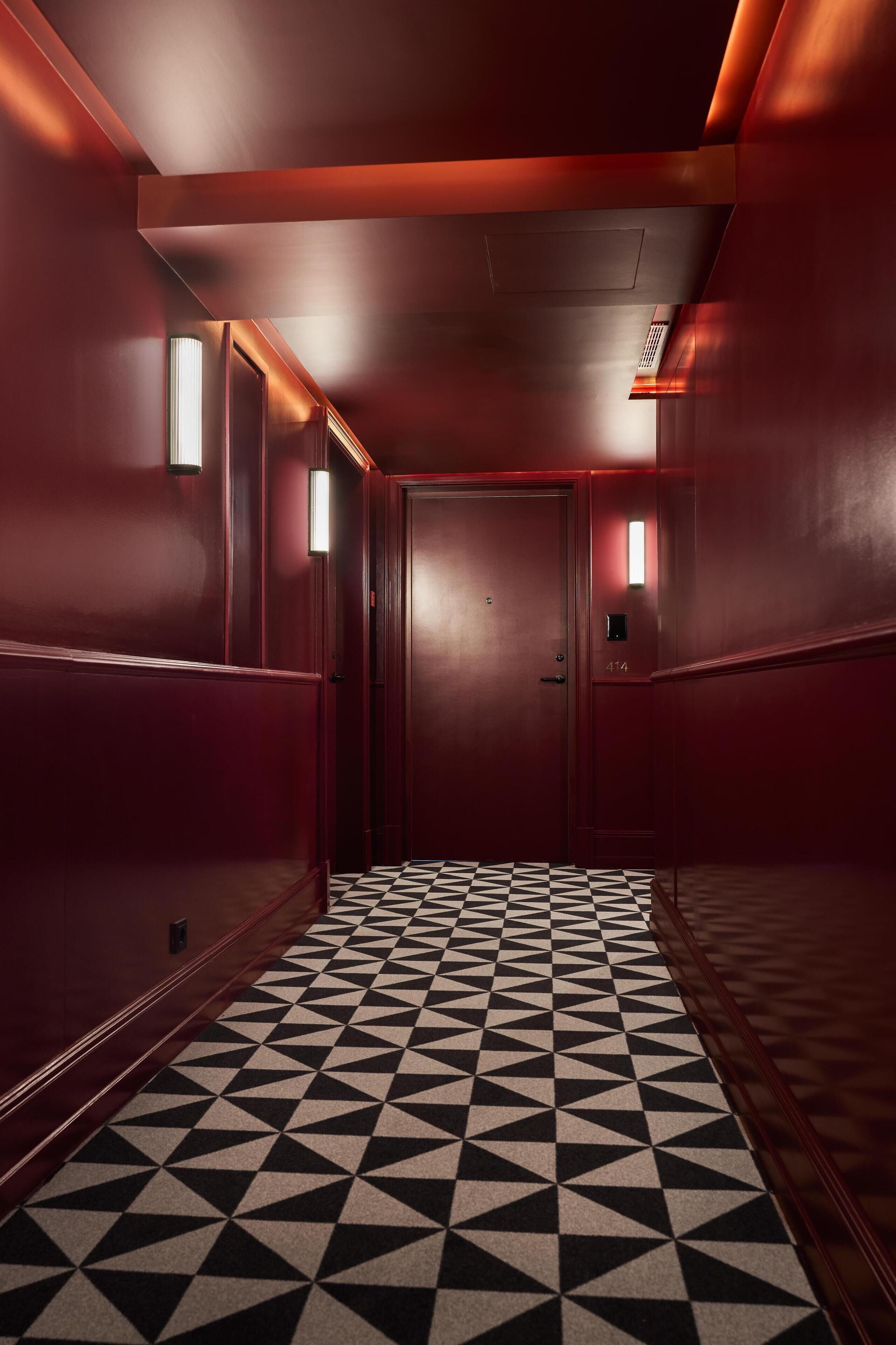 Un pasillo con piso de estampado geométrico, paredes rojas y una puerta con placa numérica.