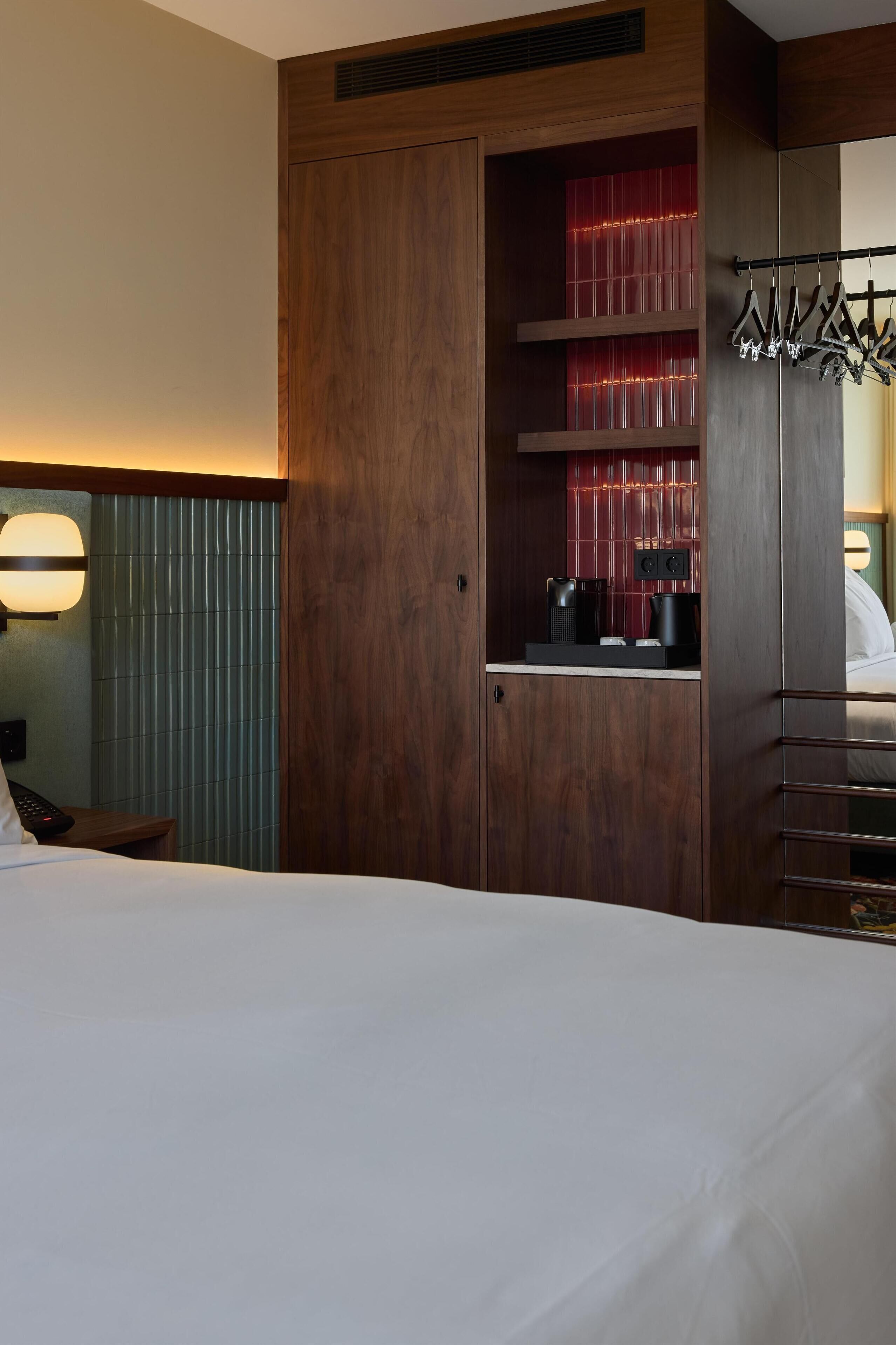 A hotel room with a large bed, a wooden wardrobe with a mirror, a red-lit shelf, and a coat rack.
