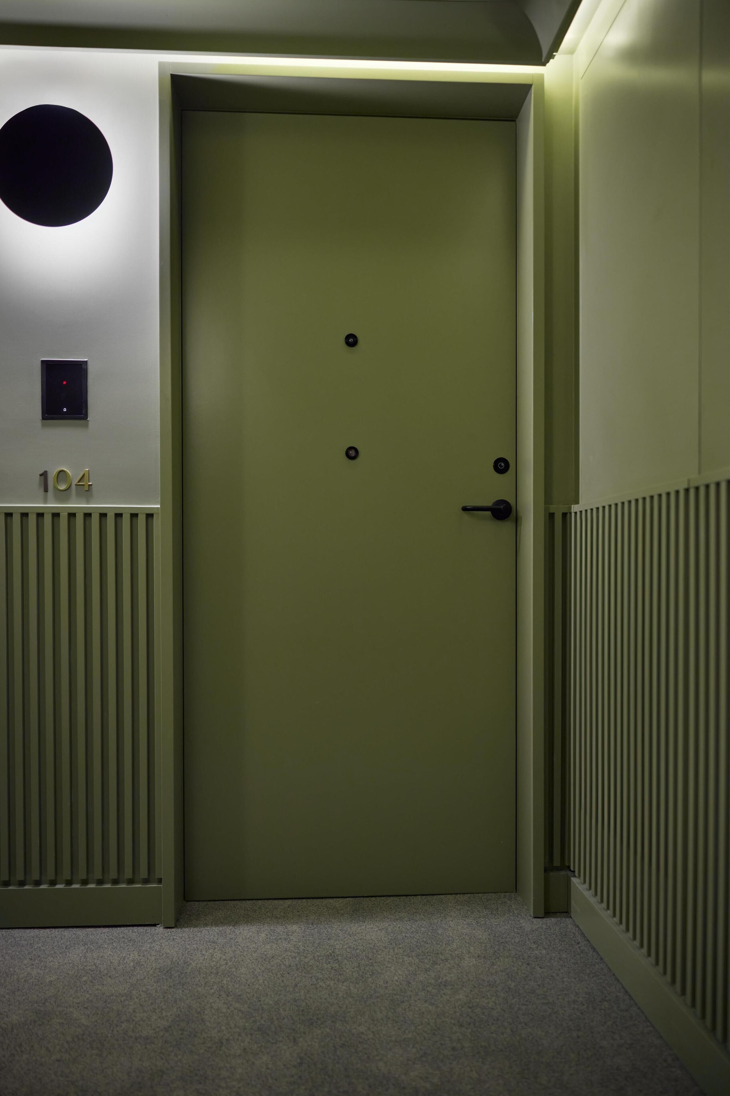 A green door with a number plate, a control panel, and a vented wall.