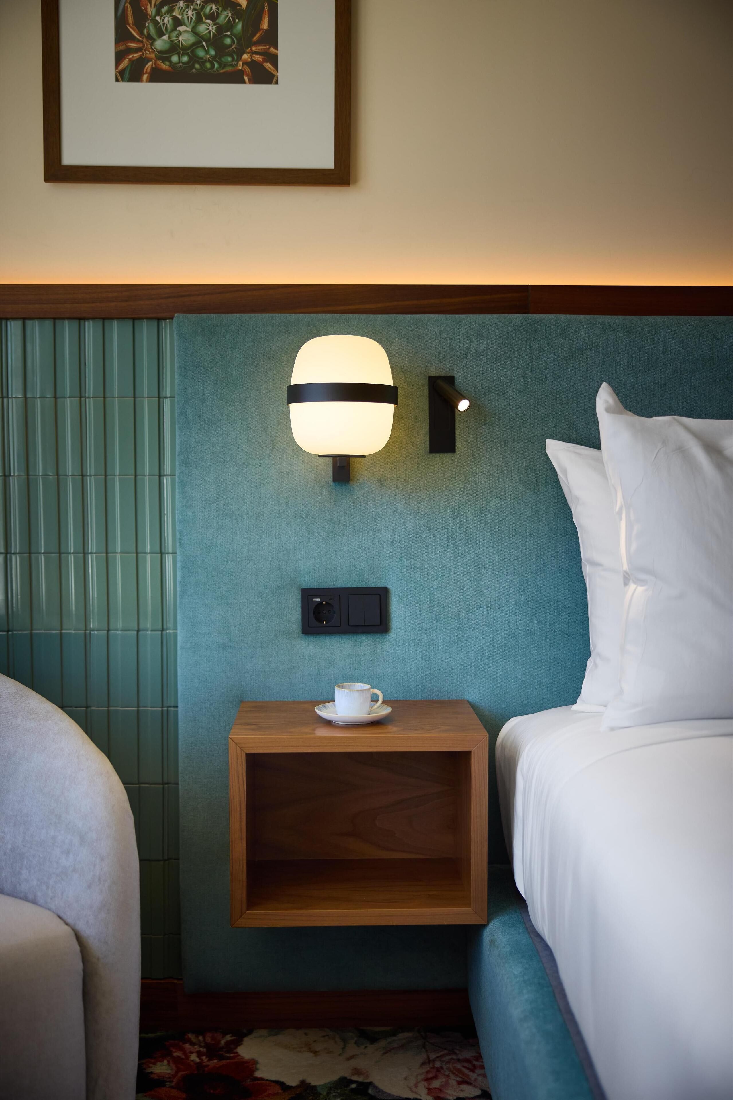 Een hotelkamer met een bed, een nachtkastje met een beker, een wandlamp en een lijst met een afbeelding.