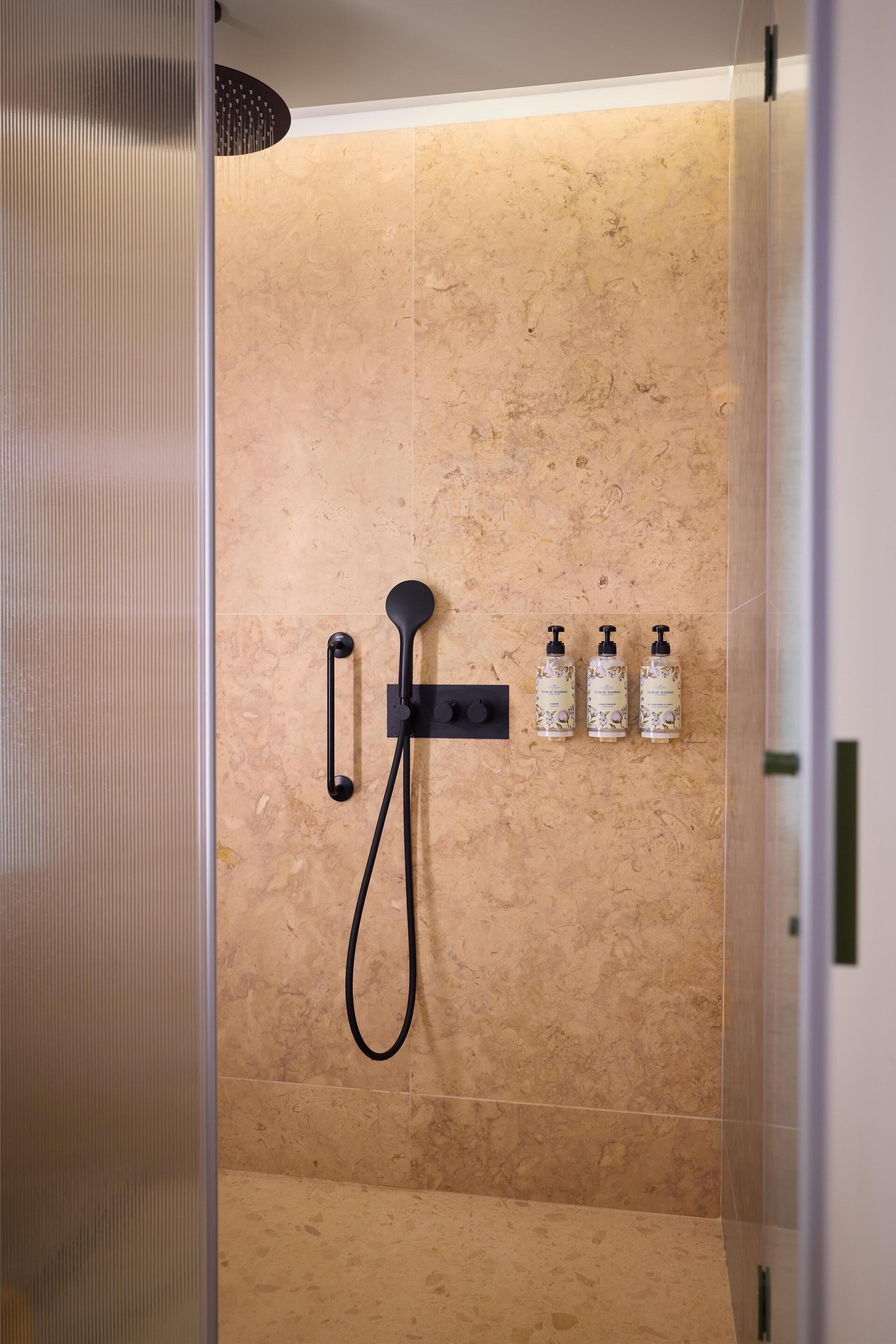 Zona de ducha con cabezal de ducha de mano, panel de control en la pared y dos botellas de champú.
