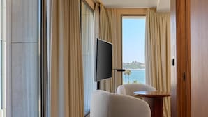 Ein modernes Hotelzimmer mit großem Fenster, einem Flachbildfernseher, einem weißen Sessel und einem runden Holztisch.