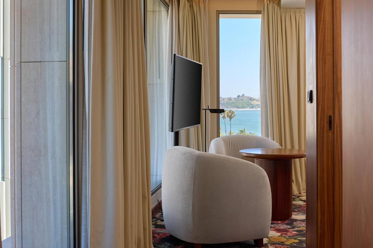 Une chambre d’hôtel moderne dotée d’une grande fenêtre, d’un téléviseur à écran plat, d’un fauteuil blanc et d’une table ronde en bois.