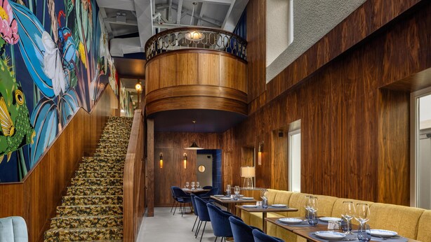 Ein modernes Interieur mit einer Holztreppe, einer Essecke mit blauen Stühlen und einem farbenfrohen Wandgemälde.